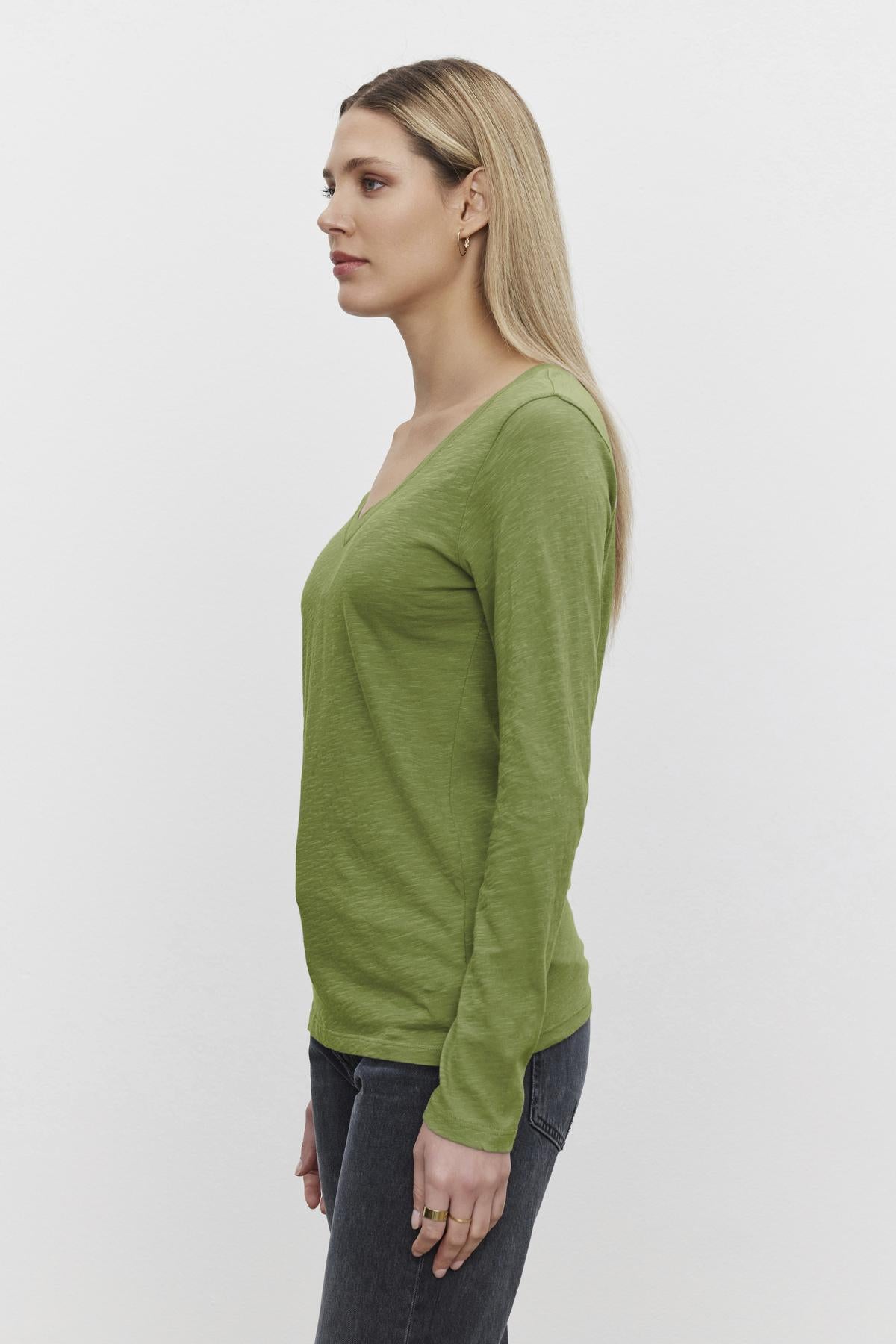   Individual with long blonde hair, wearing the Velvet by Graham & Spencer BLAIRE TEE in green and jeans, standing in profile against a white background—ideal for everyday wear. 