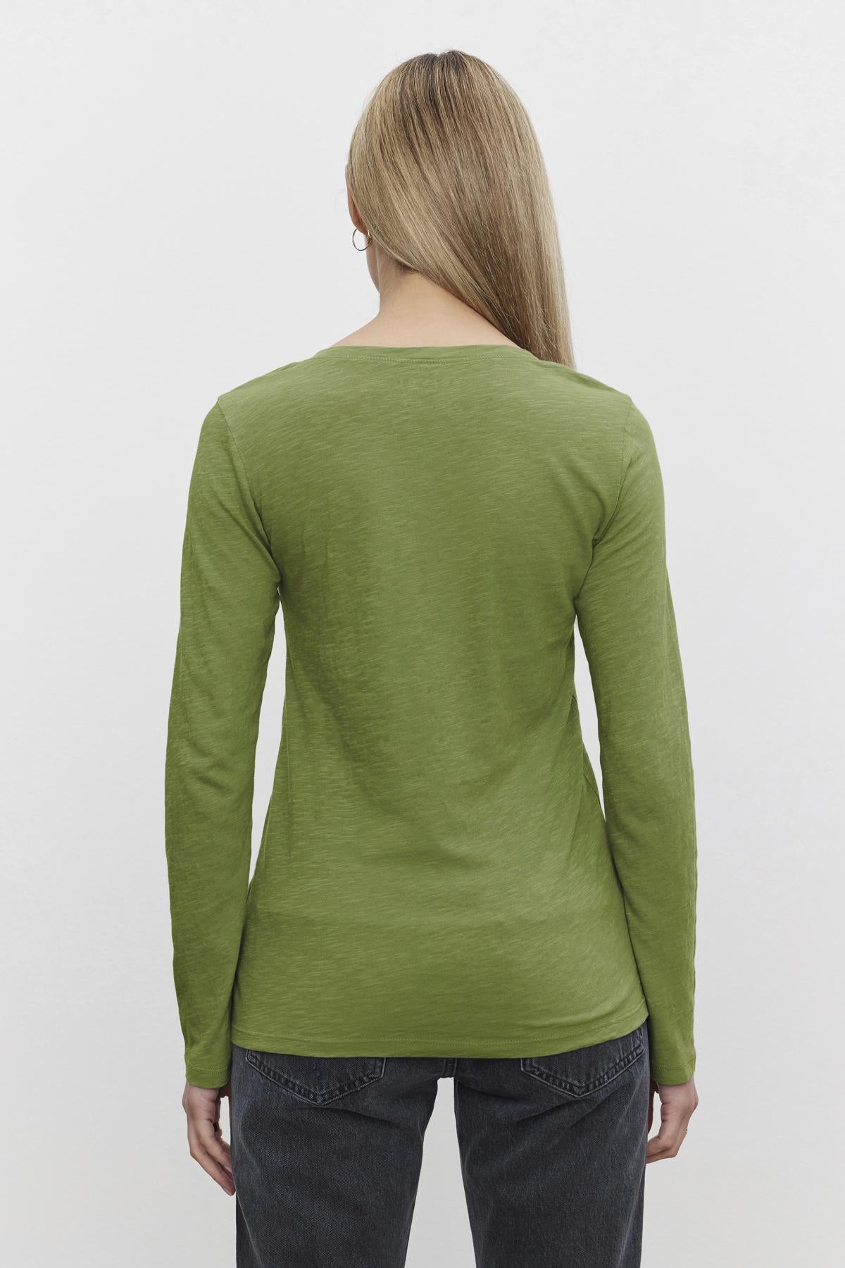   A person with long hair is seen from behind wearing a Velvet by Graham & Spencer's BLAIRE TEE, a green cotton slub shirt featuring an effortless v-neckline, paired with dark pants, against a plain white background. 