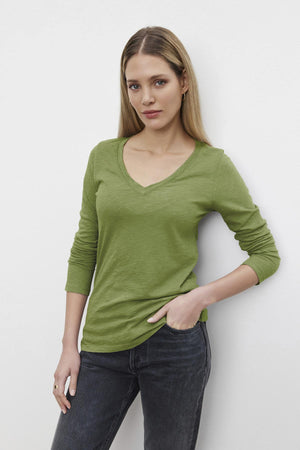 A woman with long hair poses against a white backdrop in a BLAIRE TEE by Velvet by Graham & Spencer, showcasing its textured cotton slub in green with a V-neckline. She pairs it with jeans, looking at the camera and casually placing one hand in her pocket.