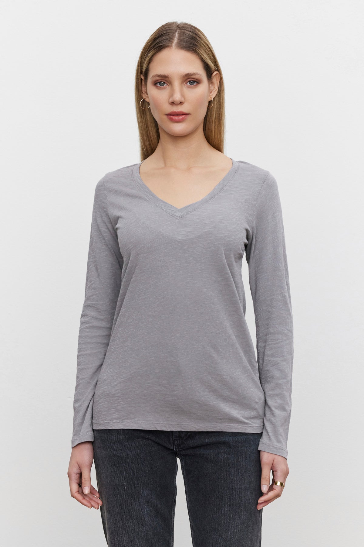   A person with long hair wearing the BLAIRE TEE from Velvet by Graham & Spencer—a gray, textured cotton slub V-neck shirt—and dark jeans stands facing forward against a plain white background. 