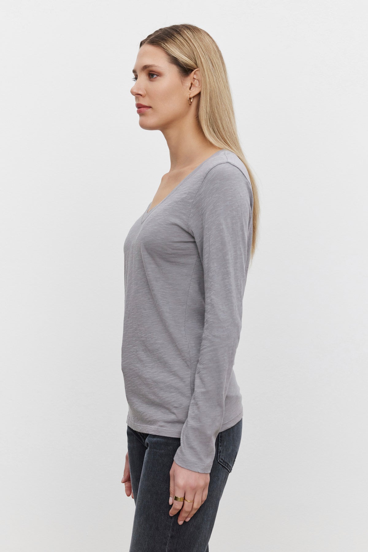   Side profile of a woman with long blonde hair, wearing the Velvet by Graham & Spencer BLAIRE TEE in gray paired with dark jeans, standing against a plain white background. 