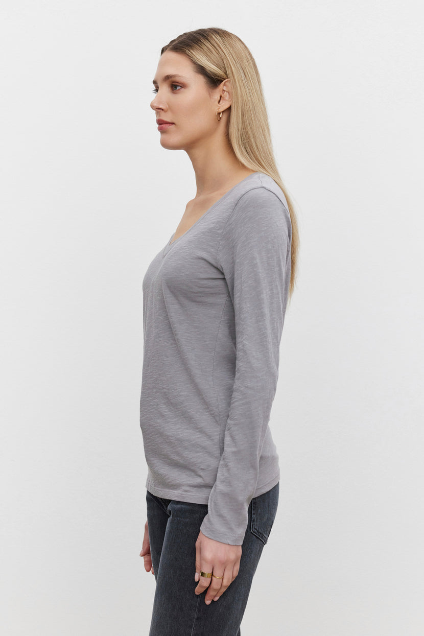 Side profile of a woman with long blonde hair, wearing the Velvet by Graham & Spencer BLAIRE TEE in gray paired with dark jeans, standing against a plain white background.