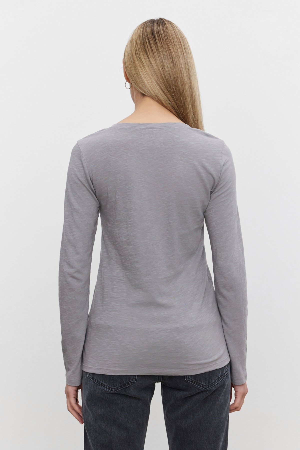   A person with long blonde hair, wearing the Velvet by Graham & Spencer's BLAIRE TEE in gray and dark jeans, is shown from the back against a plain white background. 