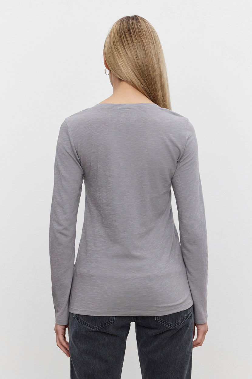 A person with long blonde hair, wearing the Velvet by Graham & Spencer's BLAIRE TEE in gray and dark jeans, is shown from the back against a plain white background.