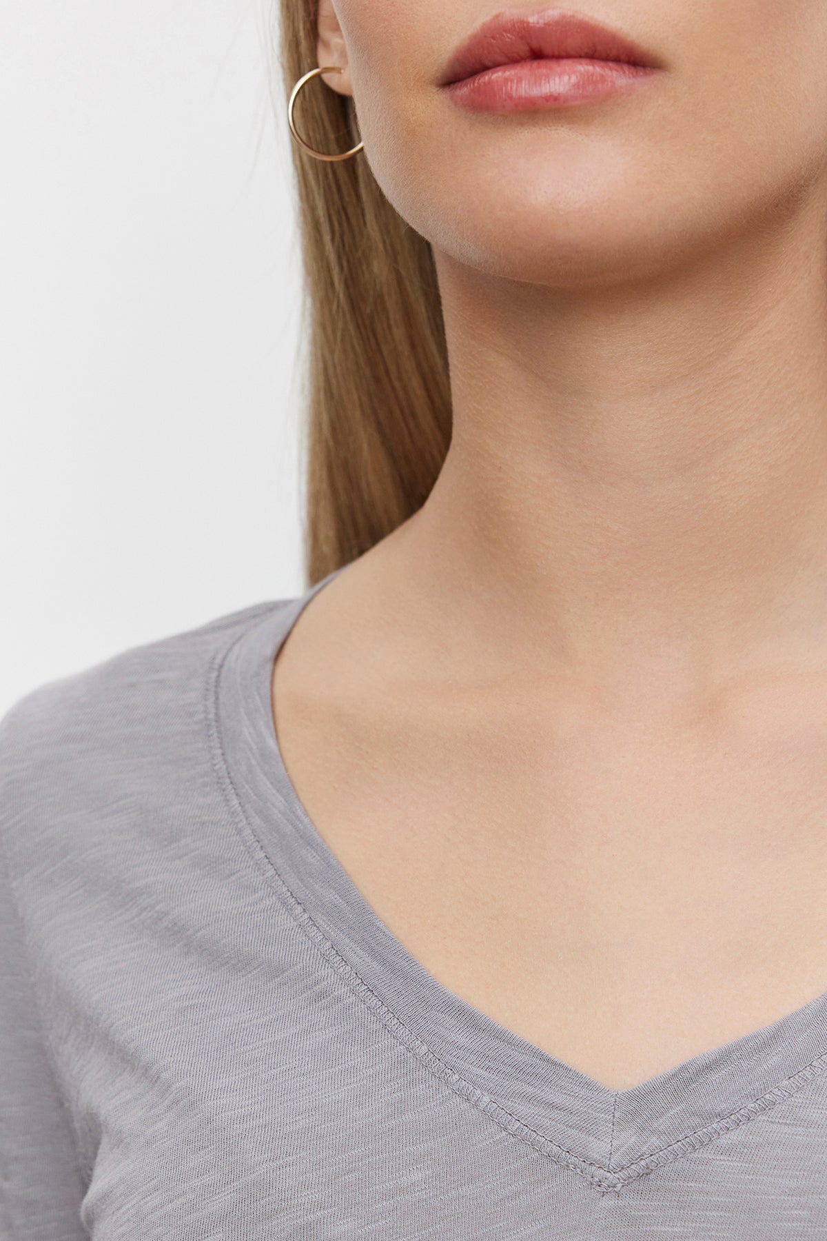   Close-up of a person wearing the BLAIRE TEE from Velvet by Graham & Spencer, a light gray textured cotton slub V-neckline shirt, and a hoop earring, with their face partially out of the frame. 