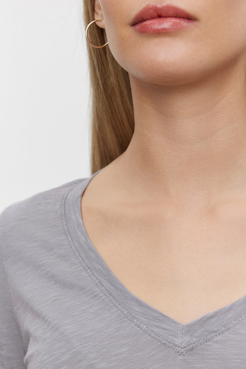 Close-up of a person wearing the BLAIRE TEE from Velvet by Graham & Spencer, a light gray textured cotton slub V-neckline shirt, and a hoop earring, with their face partially out of the frame.