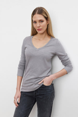 A person with long blonde hair wearing a grey Velvet by Graham & Spencer BLAIRE TEE, made of textured cotton slub with a V-neckline and long sleeves, paired with dark jeans, stands against a plain white background.