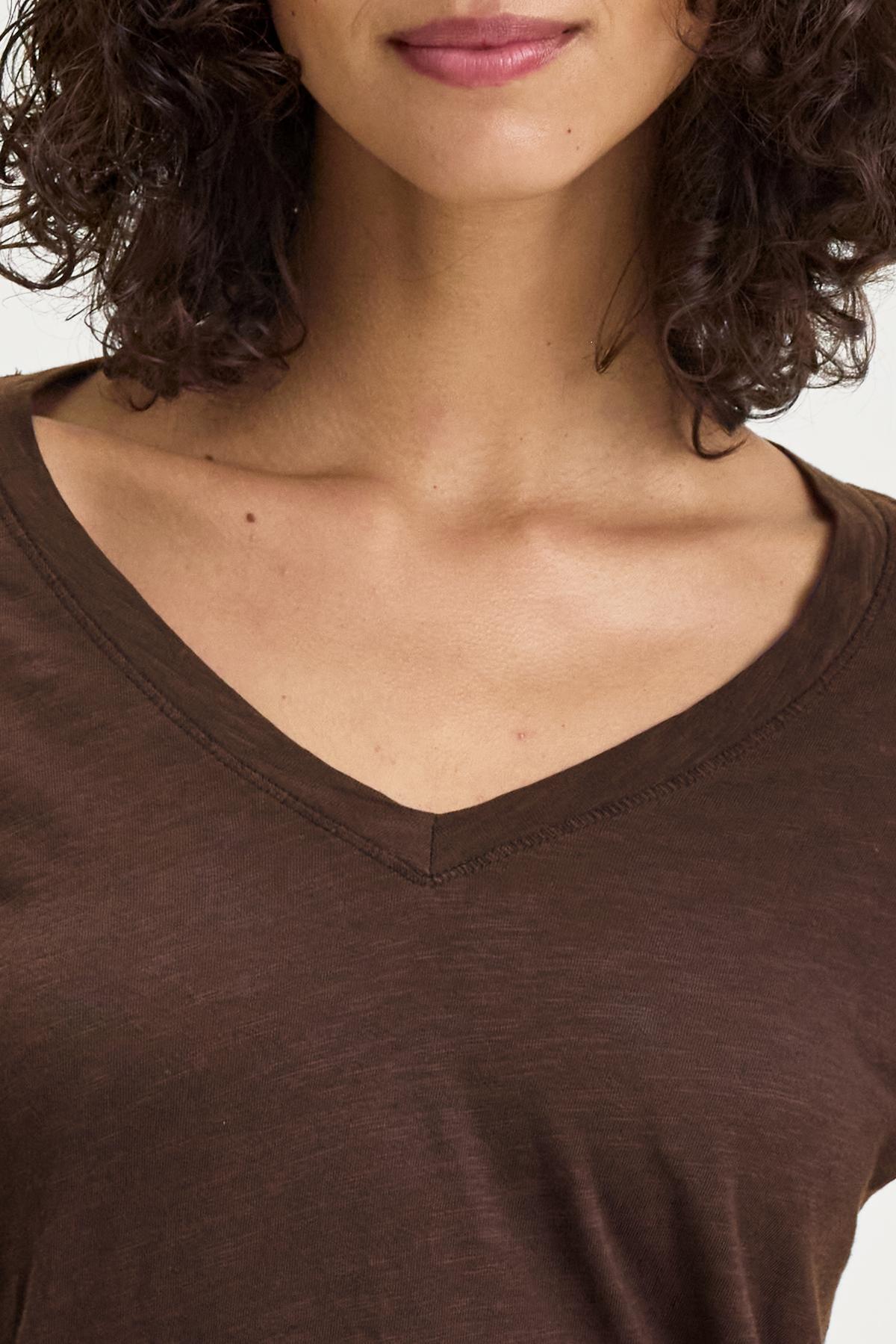   Close-up of a person with curly hair, wearing the BLAIRE TEE, a brown V-neck shirt made from textured cotton slub by Velvet by Graham & Spencer. Only the lower half of the face and upper torso are visible, showcasing comfortable everyday wear. 