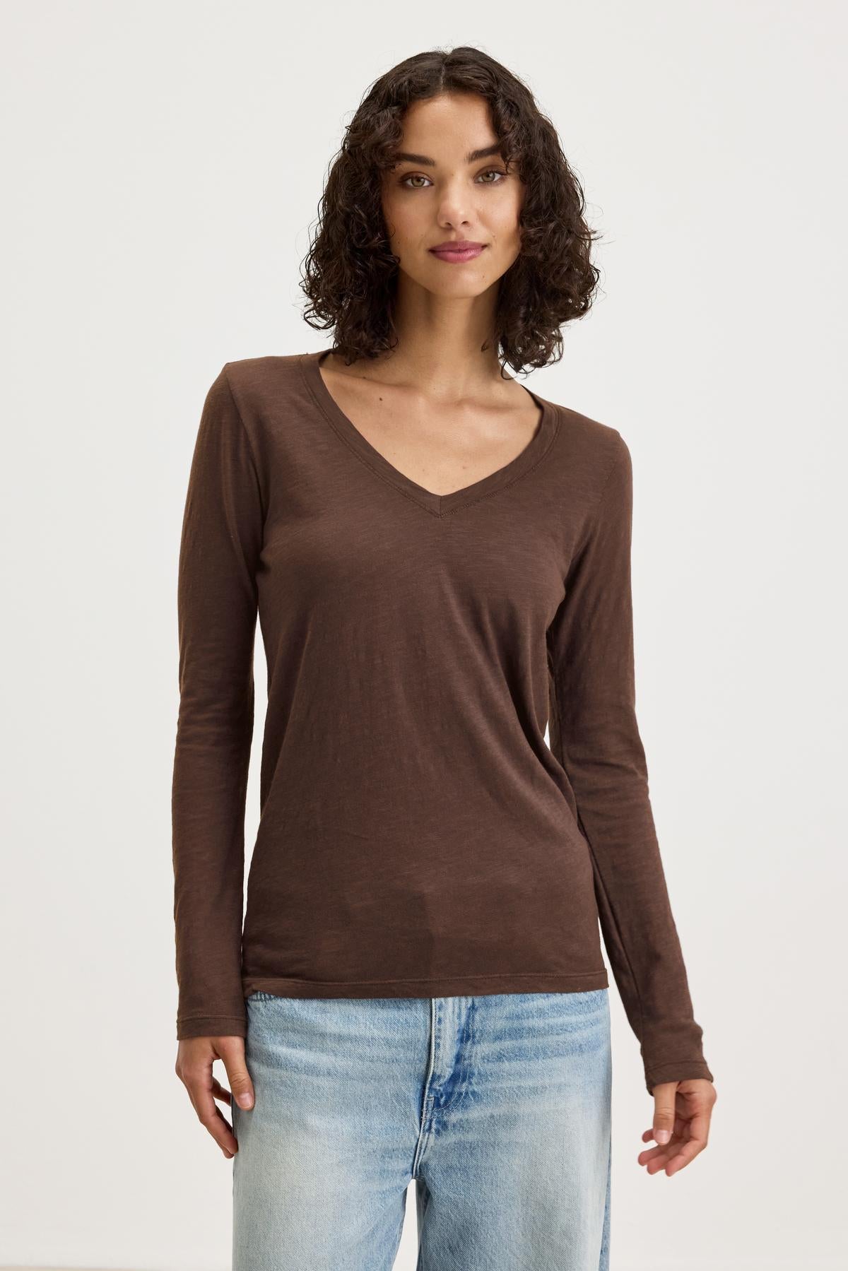   A person with curly hair is wearing a BLAIRE TEE from Velvet by Graham & Spencer, which is a brown long-sleeve V-neck shirt made of textured cotton slub, paired with light blue jeans and standing against a plain background. 