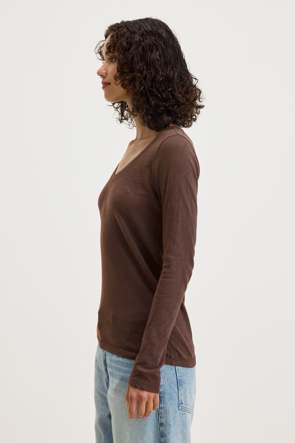   A person with curly hair is standing in profile, wearing the BLAIRE TEE in brown by Velvet by Graham & Spencer and light blue jeans. The v-neckline and textured cotton slub fabric of the shirt enhance its appeal for everyday wear. The background is plain white. 