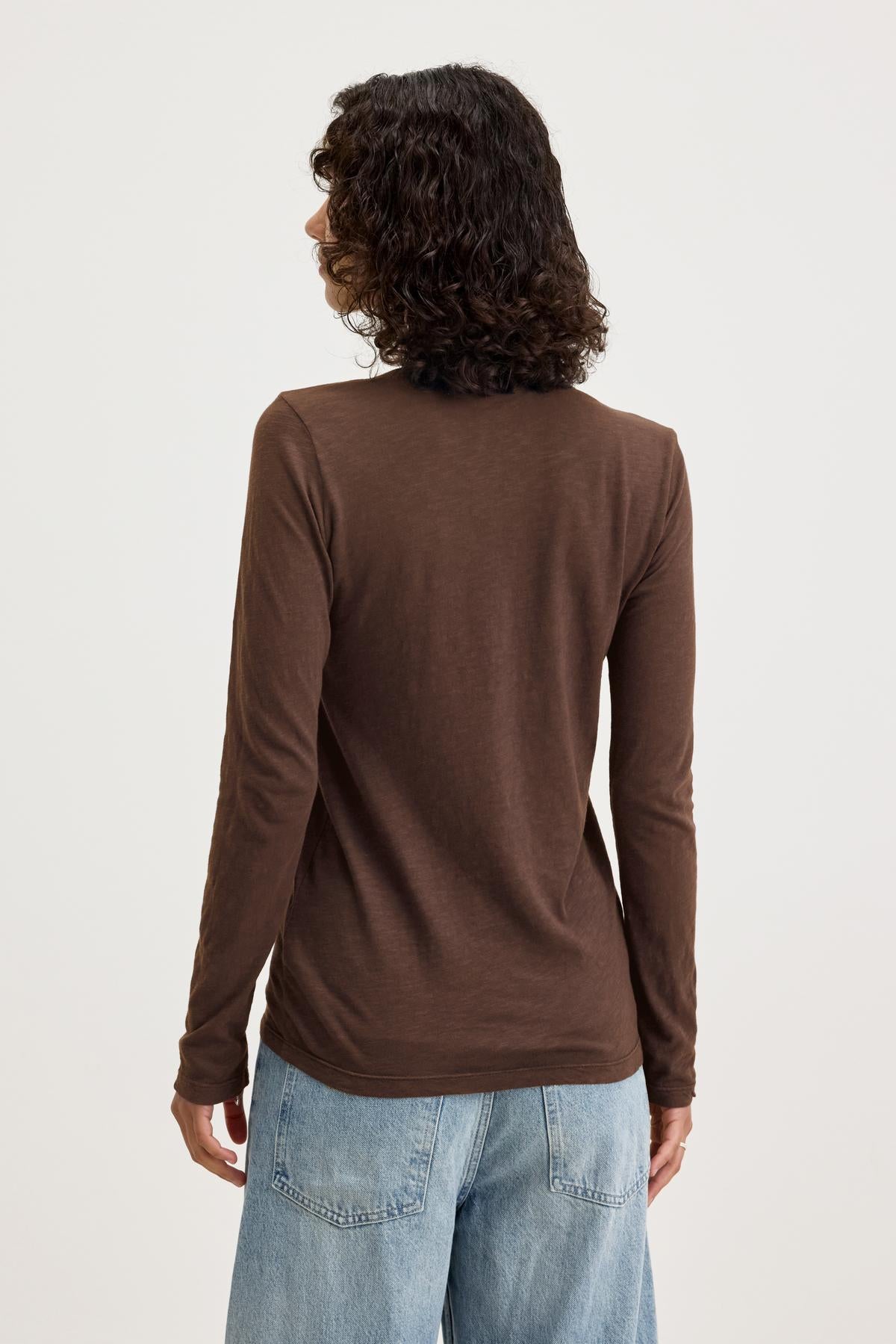   A person with curly hair is standing with their back to the camera, wearing a long-sleeved BLAIRE TEE by Velvet by Graham & Spencer in textured brown cotton slub and blue jeans. 