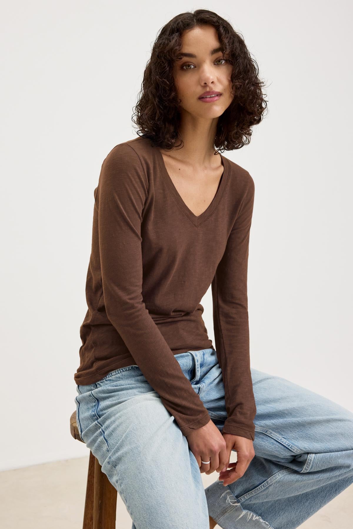   A person with curly hair, wearing a BLAIRE TEE from Velvet by Graham & Spencer and light blue jeans, sits on a stool against a plain background. 