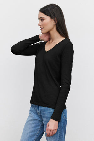 A woman with straight long hair is wearing a black V-neck long-sleeve BLAIRE TEE from Velvet by Graham & Spencer and blue jeans. She is standing sideways and looking to her right against a plain white background, epitomizing everyday wear.