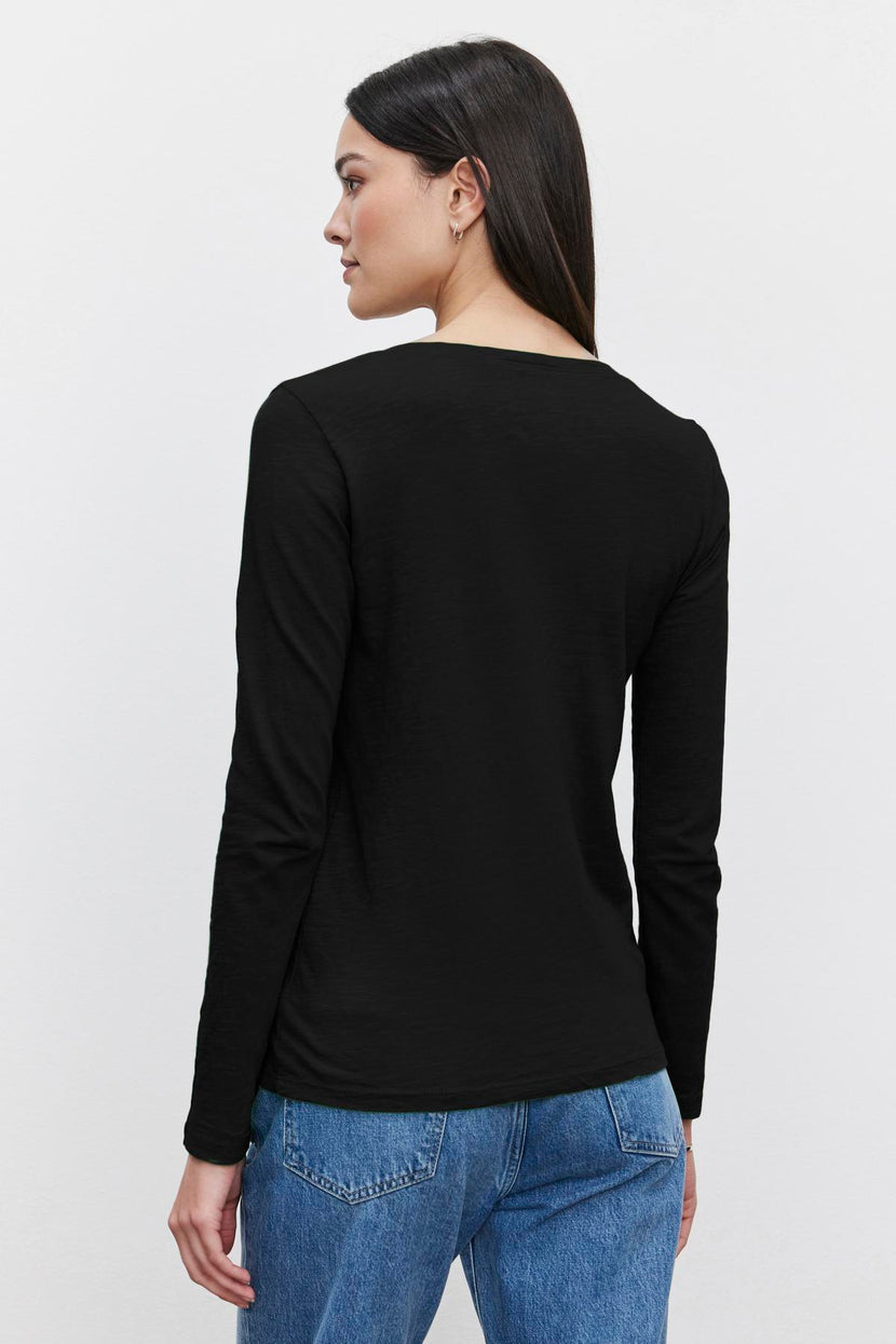 Person with long, dark hair wearing a black Velvet by Graham & Spencer BLAIRE TEE featuring a v-neckline and blue jeans, standing with their back to the camera against a plain white background.