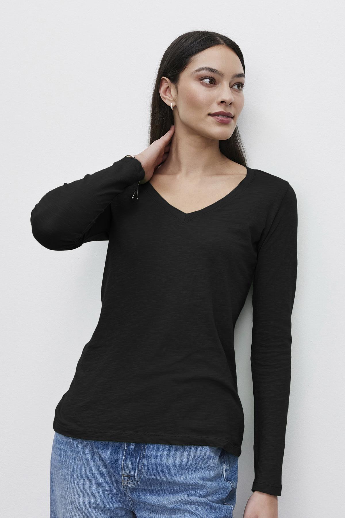   A person with long dark hair, wearing a black textured BLAIRE TEE by Velvet by Graham & Spencer and blue jeans, stands against a plain white background with one hand resting on their neck. 