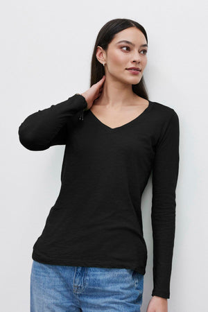 A person with long dark hair, wearing a black textured BLAIRE TEE by Velvet by Graham & Spencer and blue jeans, stands against a plain white background with one hand resting on their neck.