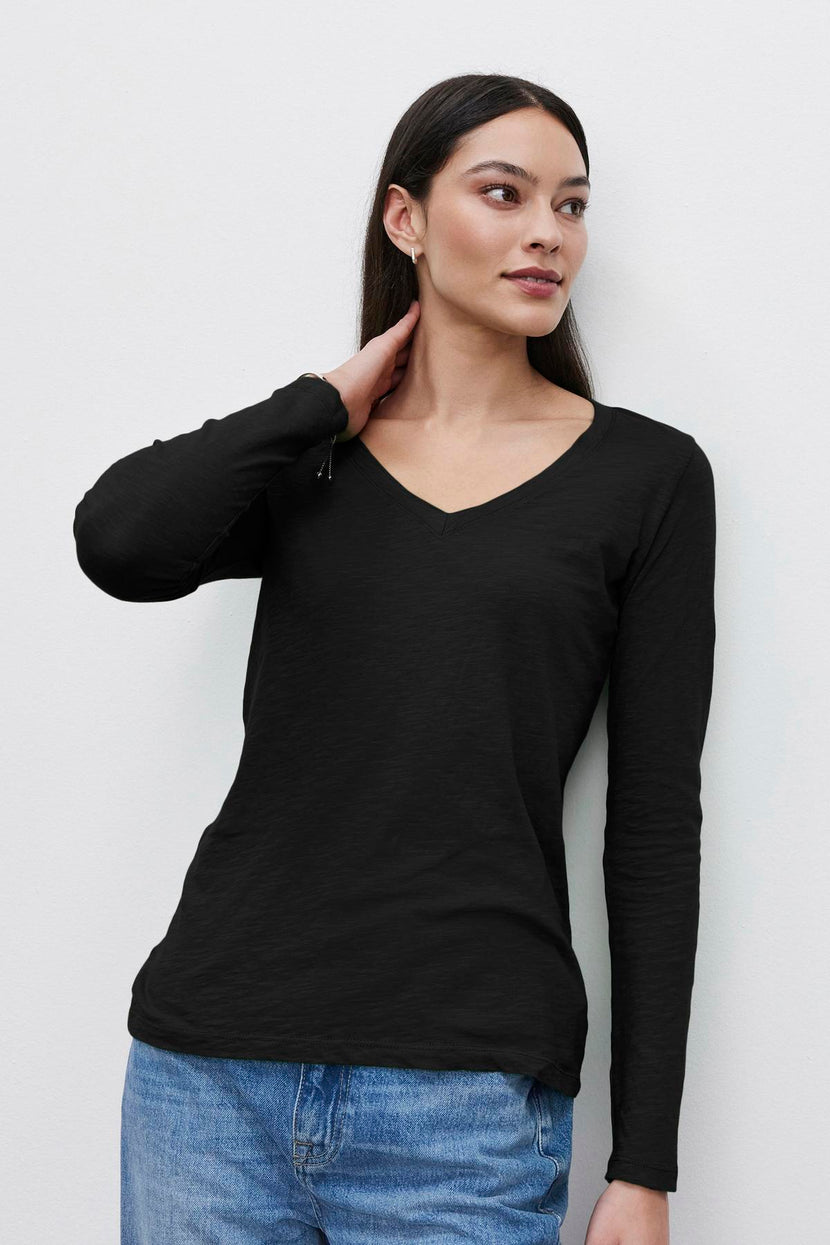 A person with long dark hair, wearing a black textured BLAIRE TEE by Velvet by Graham & Spencer and blue jeans, stands against a plain white background with one hand resting on their neck.