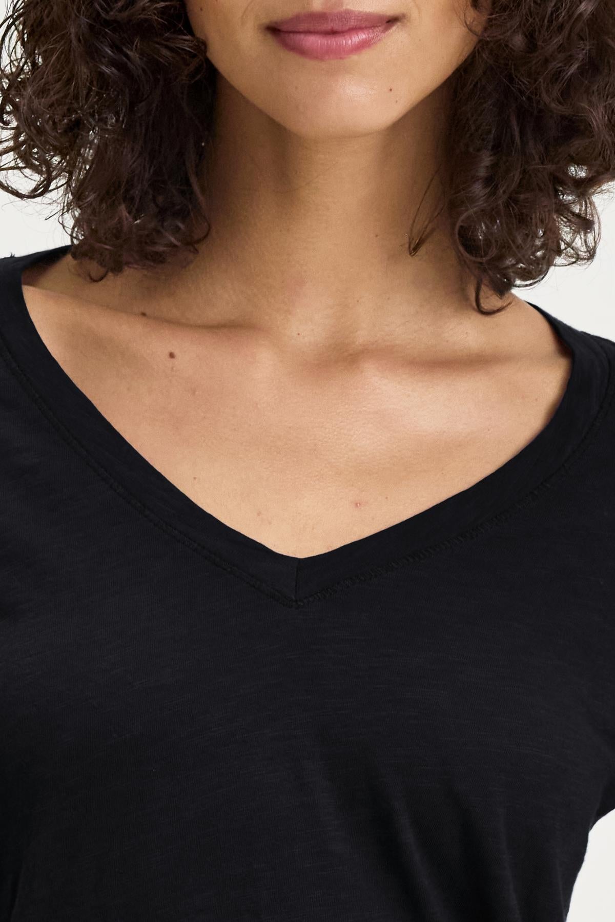   A person with curly hair wearing the BLAIRE TEE by Velvet by Graham & Spencer, featuring a black textured cotton slub and a V-neckline. The focus is on the V-neckline and shoulder area, making it perfect for everyday wear. 