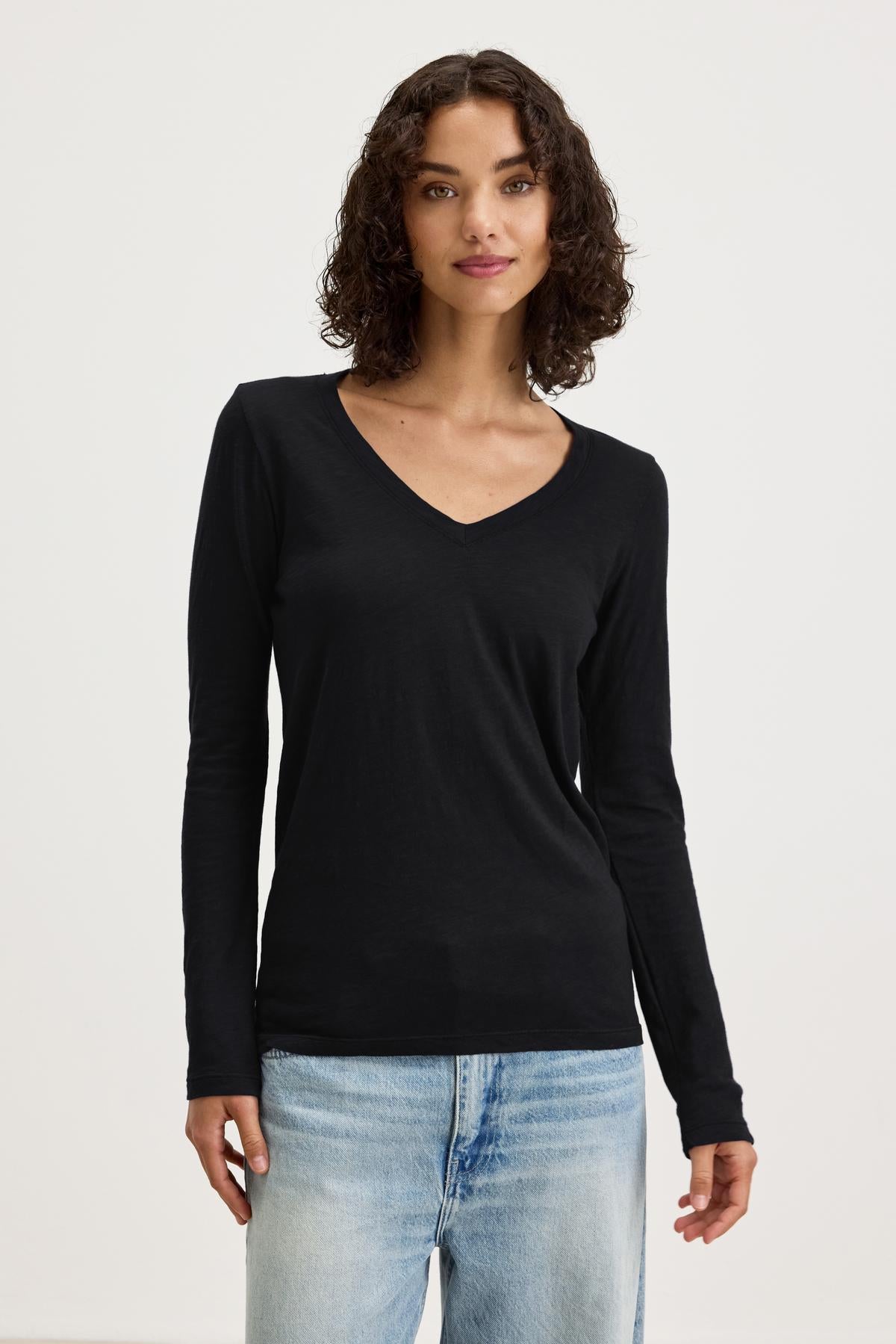   A woman with curly hair is wearing a black long-sleeve BLAIRE TEE by Velvet by Graham & Spencer and light blue jeans, perfect for everyday wear. She is standing against a plain background and looking at the camera. 