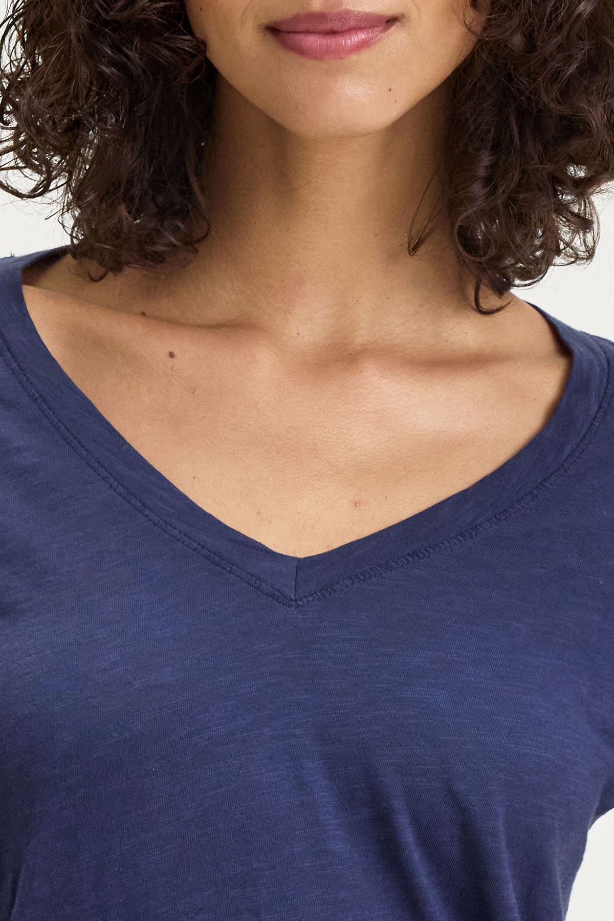   Close-up of a person wearing the BLAIRE TEE from Velvet by Graham & Spencer. The tee features a blue V-neckline and is made of textured cotton slub. The person has curly brown hair, making it perfect for everyday wear. 