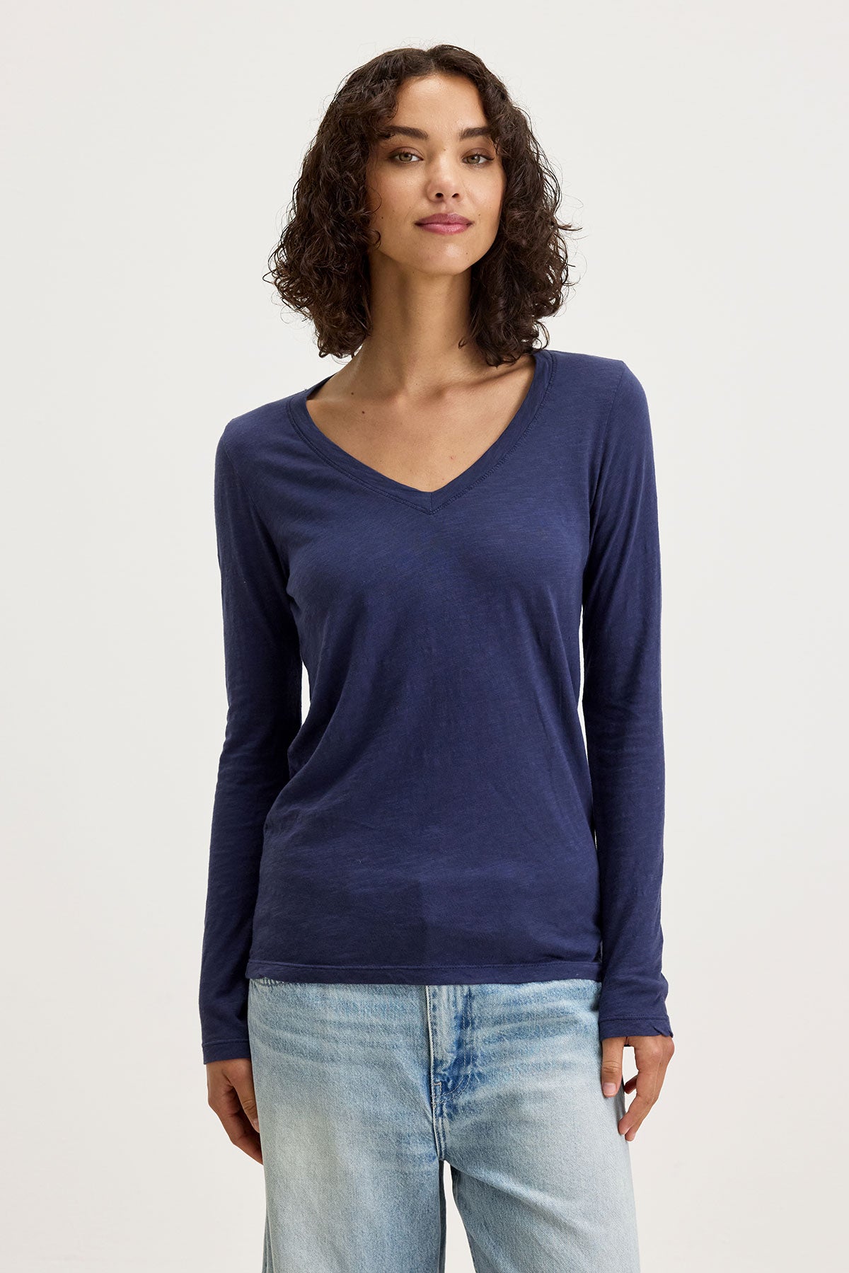   A person is standing and wearing the BLAIRE TEE, a navy blue textured cotton slub V-neckline long-sleeve shirt from Velvet by Graham & Spencer, paired with light blue jeans. They have shoulder-length curly hair and are facing the camera, embodying a perfect look for everyday wear. 