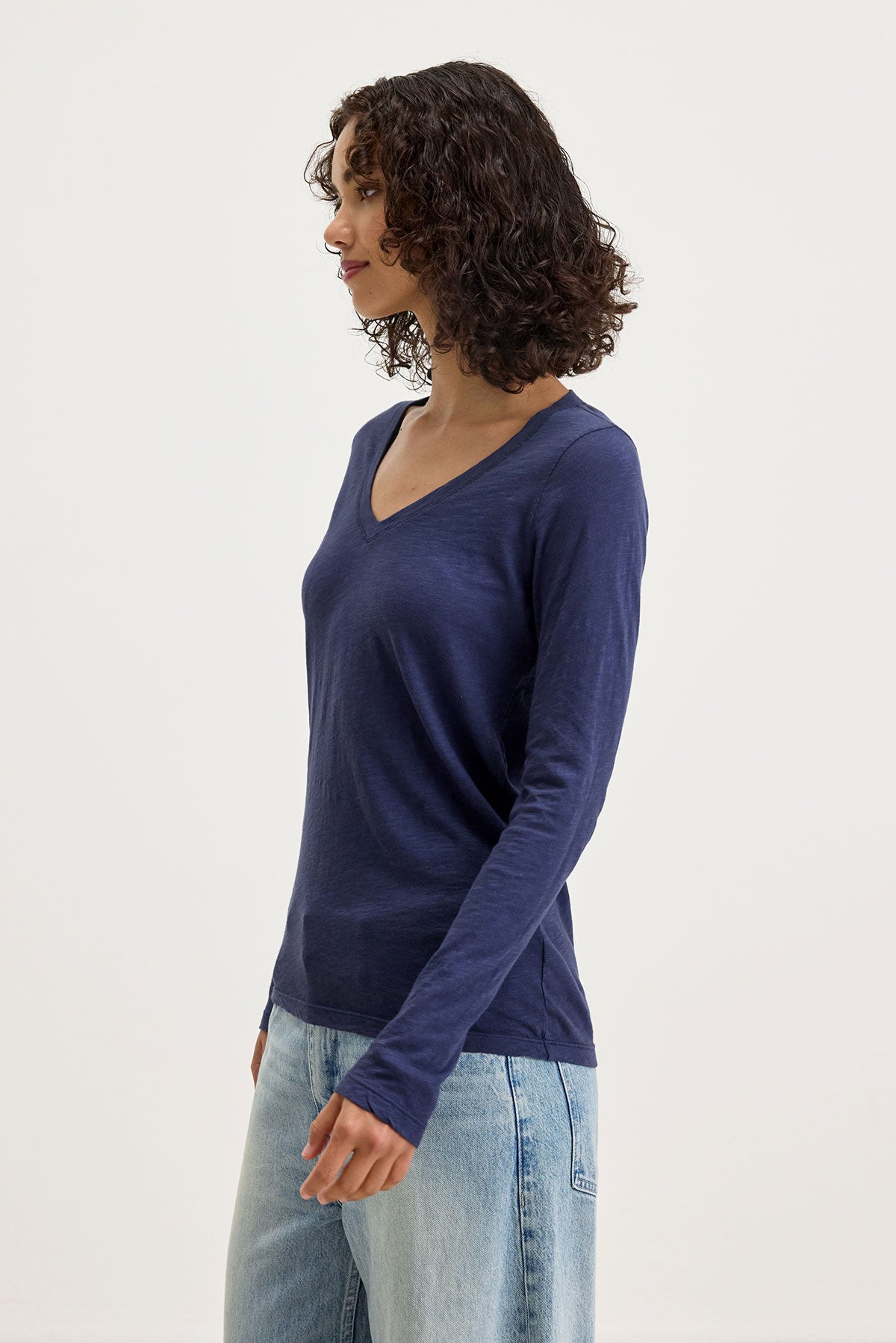   A woman with curly hair stands against a white background, wearing the Velvet by Graham & Spencer BLAIRE TEE—an elegant navy blue textured cotton slub long-sleeved V-neck shirt—and light blue jeans, making it perfect for everyday wear. 