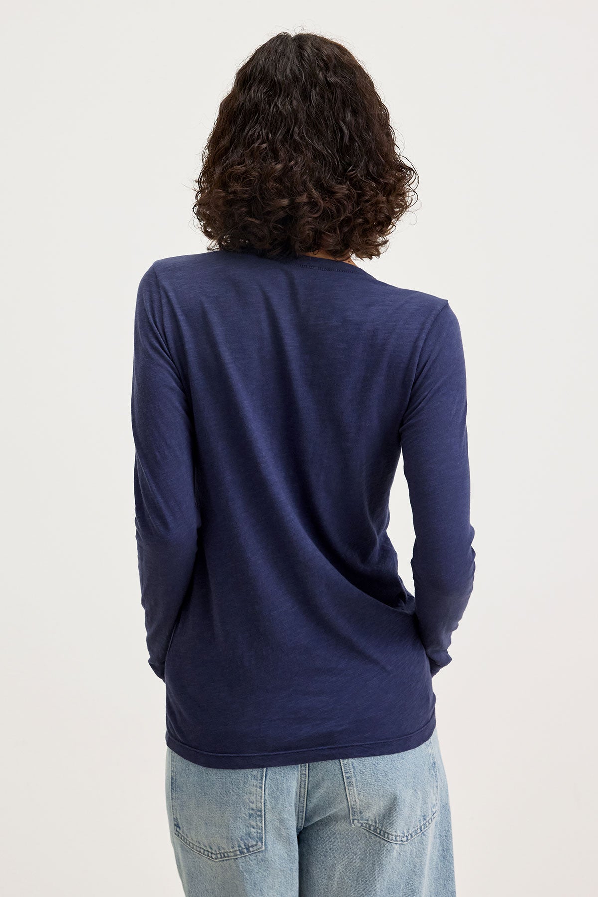   A person with curly hair, wearing a Velvet by Graham & Spencer BLAIRE TEE shirt in navy made of textured cotton slub and light blue jeans, is shown from the back against a plain background. 