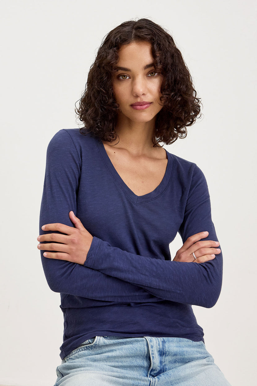 Person with curly dark hair wearing the BLAIRE TEE by Velvet by Graham & Spencer, a long-sleeve dark blue v-neck shirt, paired with light blue jeans, crossing their arms and standing against a plain light background.