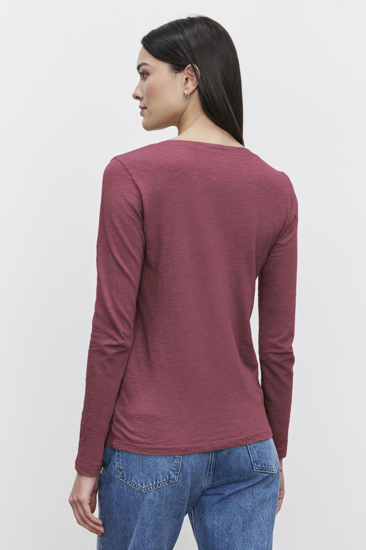   A person with long hair is wearing the maroon BLAIRE TEE from Velvet by Graham & Spencer and blue jeans, facing away from the camera against a plain white background. 