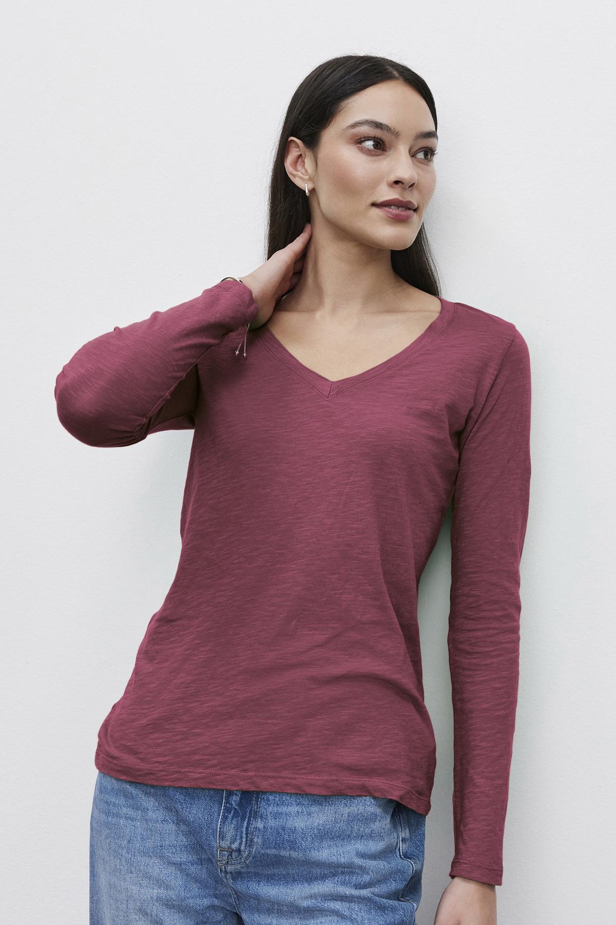   Wearing a maroon BLAIRE TEE by Velvet by Graham & Spencer, featuring a long-sleeve V-neckline crafted from textured cotton slub, a person stands against a light background, effortlessly paired with blue jeans for everyday wear. 