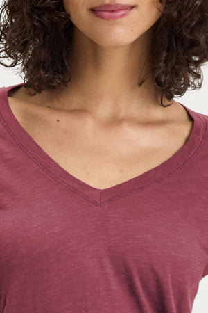 A person is seen in everyday wear, donning the Velvet by Graham & Spencer BLAIRE TEE shirt with a maroon V-neckline made from textured cotton slub, gracefully showcasing the collarbone and complemented by their curly hair.