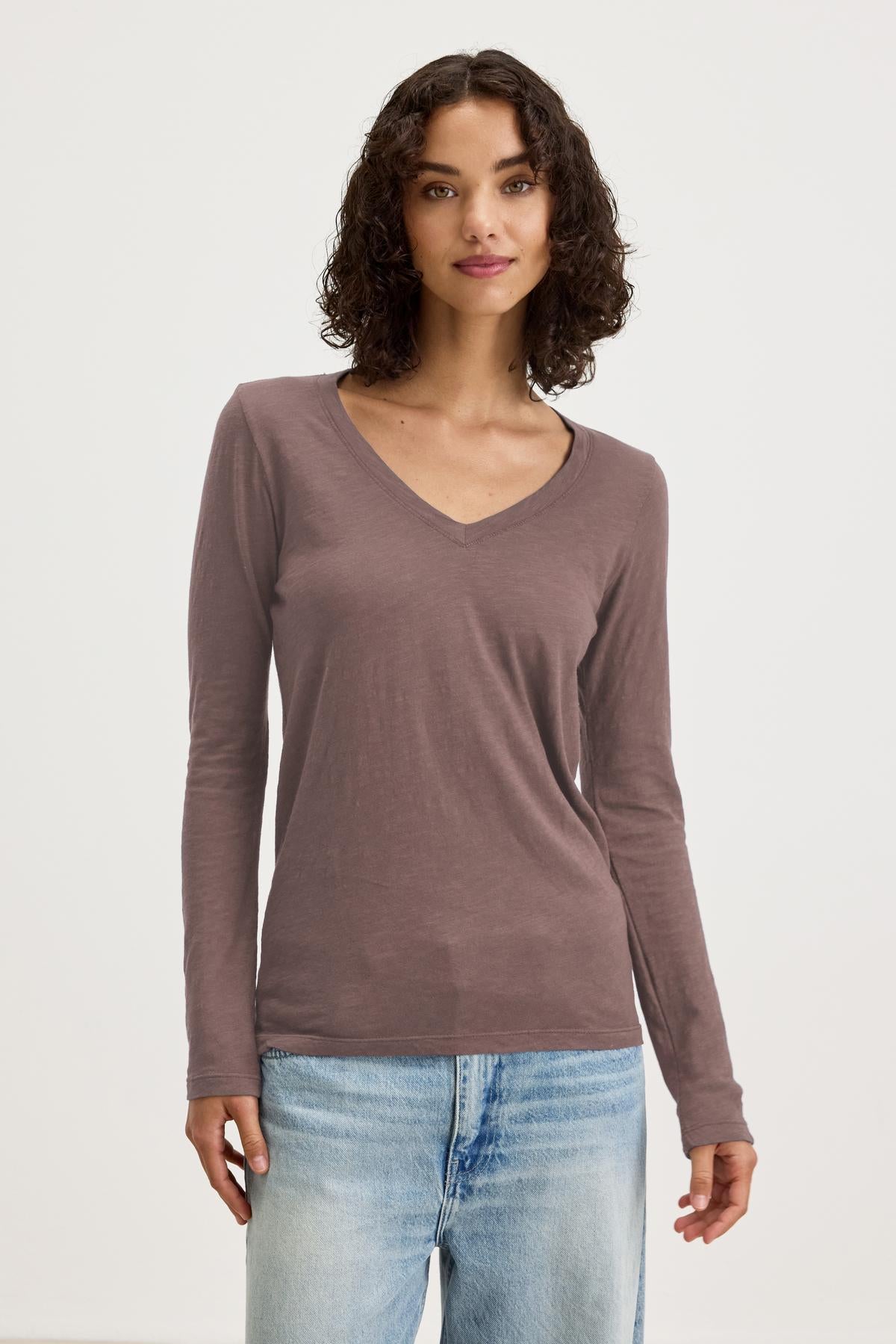   A person wears the BLAIRE TEE by Velvet by Graham & Spencer, a long-sleeve shirt with a V-neckline in earthy brown cotton slub, paired with light blue jeans and set against a plain white backdrop, ideal for everyday wear. 