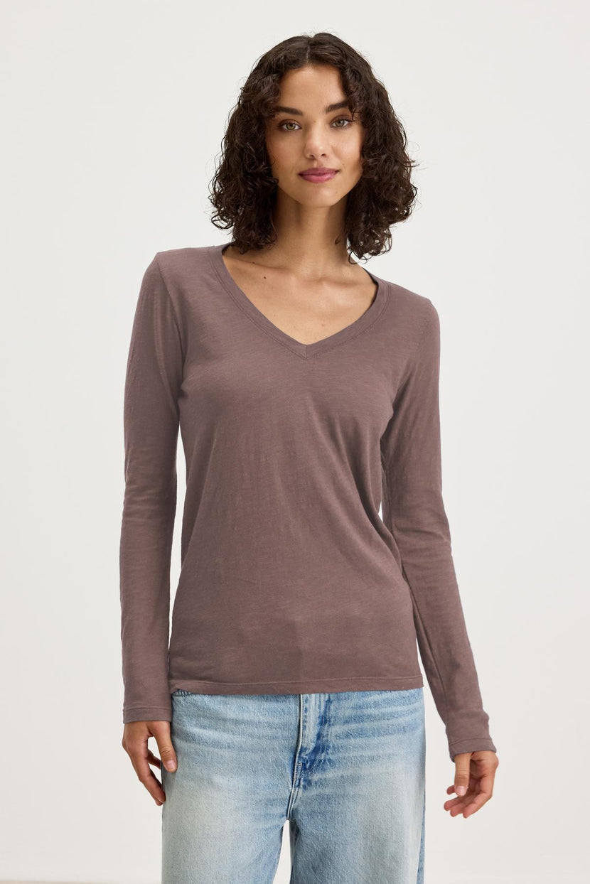 A person wears the BLAIRE TEE by Velvet by Graham & Spencer, a long-sleeve shirt with a V-neckline in earthy brown cotton slub, paired with light blue jeans and set against a plain white backdrop, ideal for everyday wear.