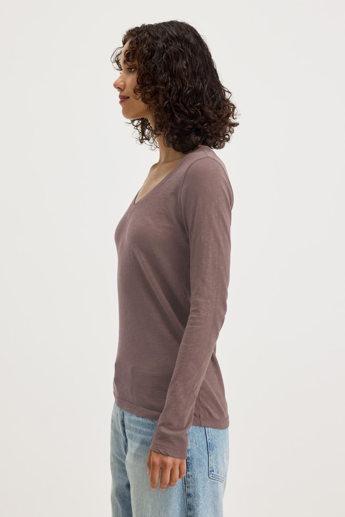   A person with curly hair is wearing Velvet by Graham & Spencer's BLAIRE TEE, a long-sleeve brown shirt with a v-neckline, along with blue jeans, standing sideways against a plain background. The outfit epitomizes everyday wear, combining comfort and style in cotton slub fabric. 