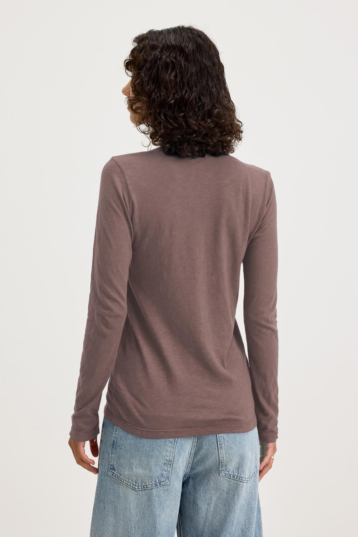   A person with curly hair, dressed in an everyday ensemble featuring the BLAIRE TEE from Velvet by Graham & Spencer and a pair of jeans. They are standing with their back to the camera, showcasing the outfit's casual elegance. 