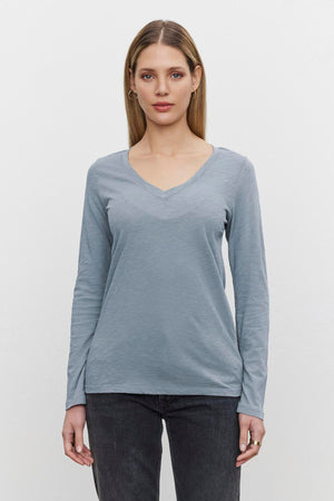 A person with long hair is wearing a light blue BLAIRE TEE from Velvet by Graham & Spencer and dark jeans, standing against a plain white background.