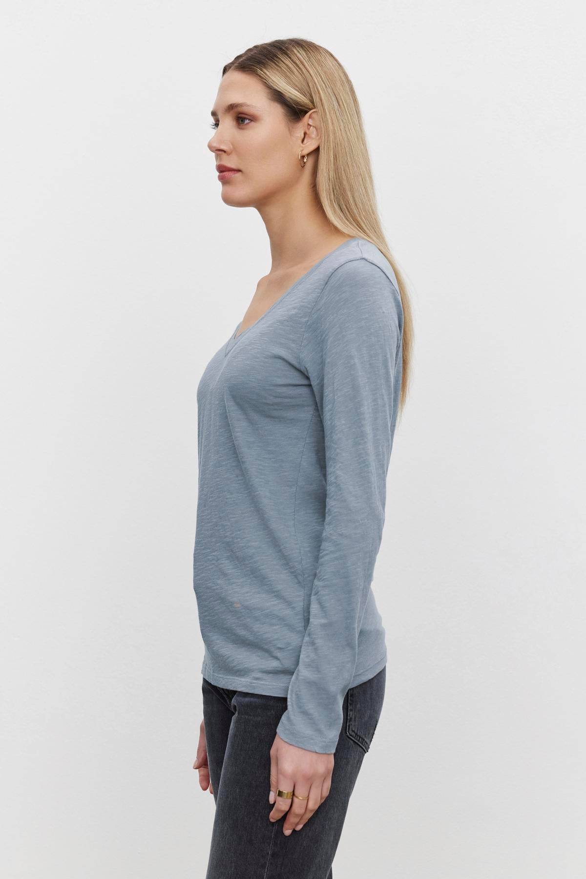   A woman with long blond hair stands in profile against a plain background, wearing the light blue V-neckline long-sleeve BLAIRE TEE by Velvet by Graham & Spencer and dark pants, perfect for everyday wear. 