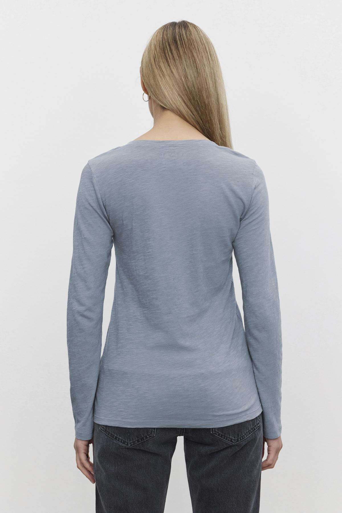   A person with long hair stands with their back to the camera, wearing a light blue BLAIRE TEE by Velvet by Graham & Spencer along with dark jeans. 