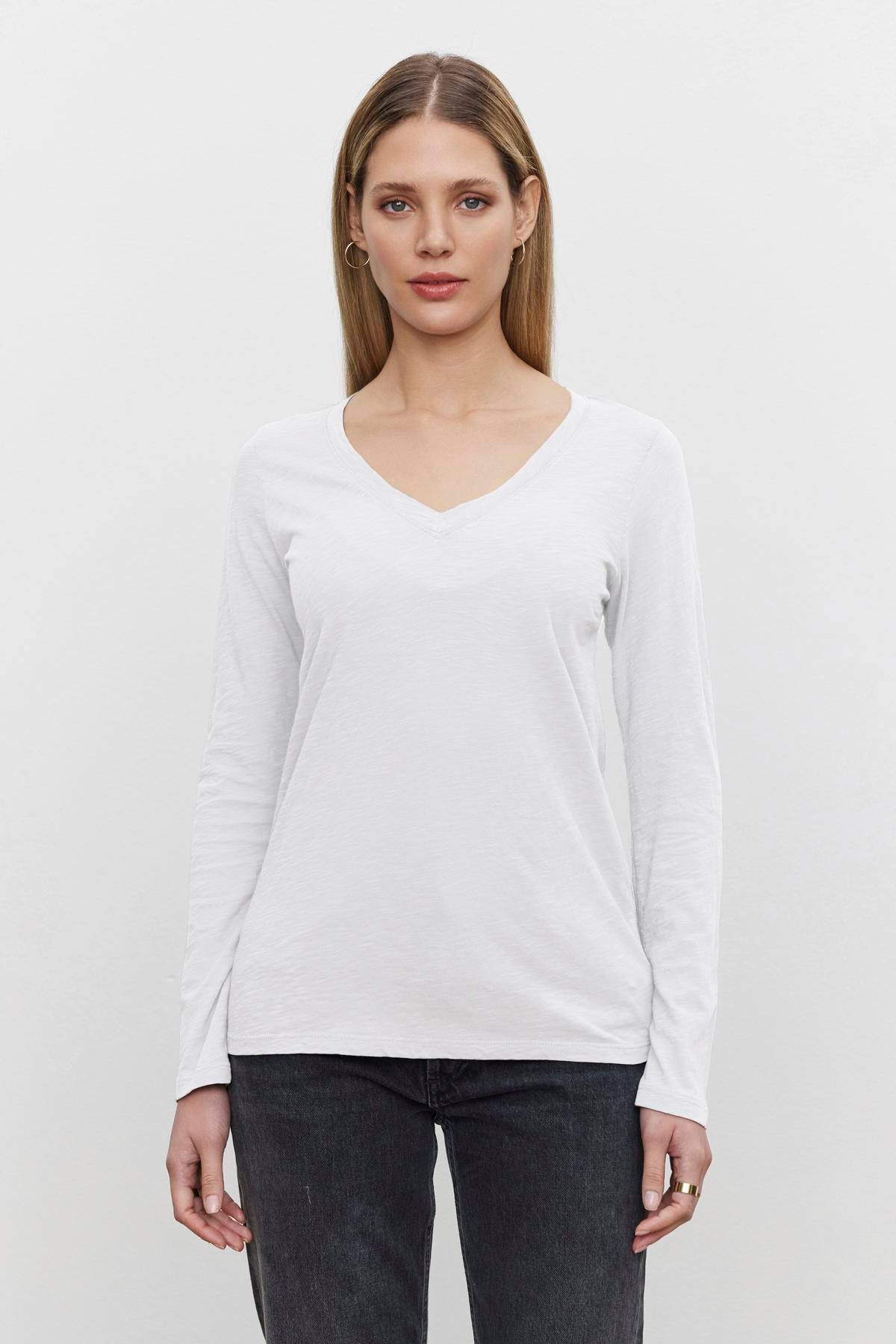   A person with long hair wearing a white BLAIRE TEE long-sleeve shirt made from textured cotton slub by Velvet by Graham & Spencer and dark jeans stands against a plain white background. 