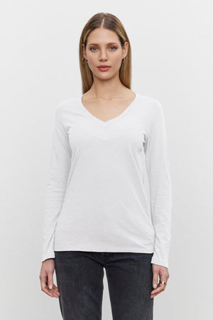 A person with long hair wearing a white BLAIRE TEE long-sleeve shirt made from textured cotton slub by Velvet by Graham & Spencer and dark jeans stands against a plain white background.