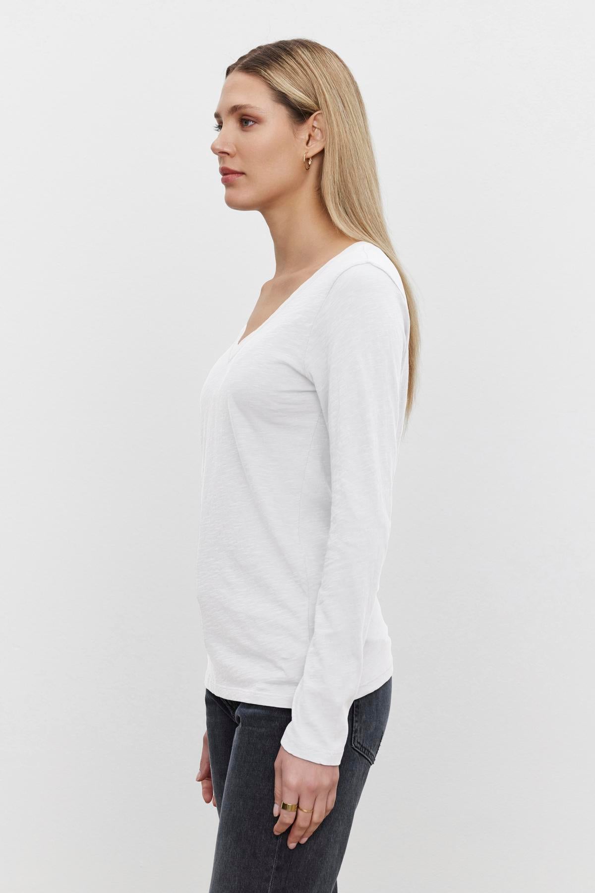 A person with long blonde hair stands in profile, wearing a white textured BLAIRE TEE by Velvet by Graham & Spencer with a v-neckline and dark jeans against a plain white background.-37377300103361