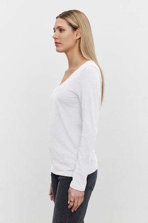 A person with long blonde hair stands in profile, wearing a white textured BLAIRE TEE by Velvet by Graham & Spencer with a v-neckline and dark jeans against a plain white background.
