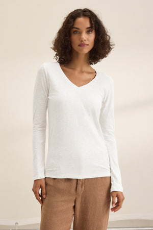 A person stands against a plain background wearing the BLAIRE TEE by Velvet by Graham & Spencer, a white cotton slub V-neck long sleeve top, paired with light brown pants.