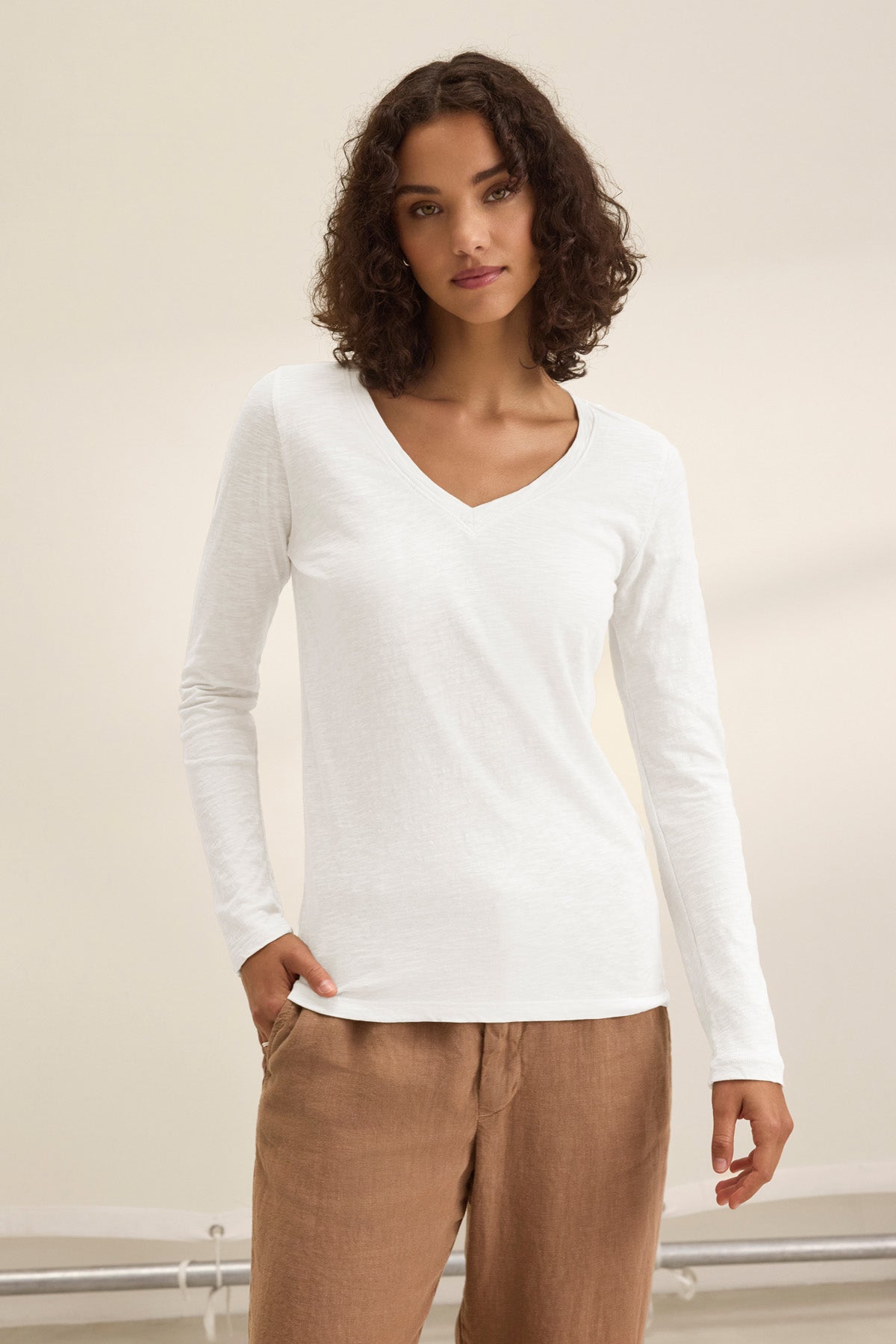 A person with curly hair models the Velvet by Graham & Spencer BLAIRE TEE—a white cotton slub V-neck long-sleeve top—paired with light brown pants, standing against a plain light background.-41476532994241