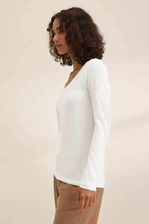 A person with curly brown hair wears the BLAIRE TEE by Velvet by Graham & Spencer—a white cotton slub, V-neck, long-sleeve top—paired with light brown pants, standing sideways against a plain beige background.