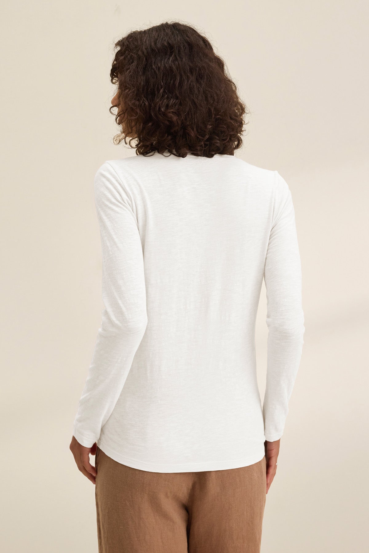 A person with curly hair is seen from the back, wearing the BLAIRE TEE by Velvet by Graham & Spencer— a long sleeve slub cotton v-neck — paired with light brown pants, standing against a plain backdrop.