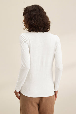 A person with curly hair is seen from the back, wearing the BLAIRE TEE by Velvet by Graham & Spencer— a long sleeve slub cotton v-neck — paired with light brown pants, standing against a plain backdrop.