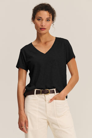 A woman stands facing forward, wearing the ELISHA TEE by Velvet by Graham & Spencer—a black cotton slub V-neck—tucked into high-waisted white pants with a dark belt against a plain background.
