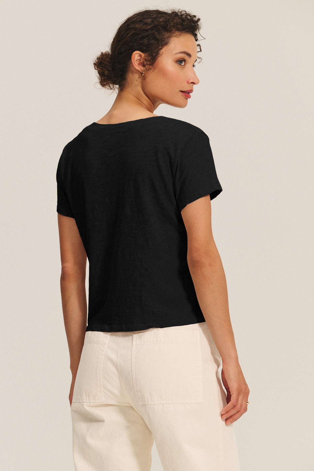 A woman stands facing away, wearing the ELISHA TEE by Velvet by Graham & Spencer—a black short-sleeve cotton slub top—with high-waisted white pants against a plain light background.