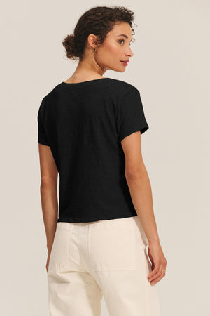 A woman stands facing away, wearing the ELISHA TEE by Velvet by Graham & Spencer—a black short-sleeve cotton slub top—with high-waisted white pants against a plain light background.